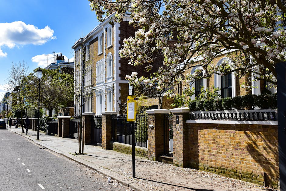 View of a residential street lined with brick and stucco houses, featuring white window frames and decorative architectural details. Several trees, some in bloom with white flowers, line the sidewalk, which has a low brick wall and black wrought iron fencing in front of the properties. A yellow bus stop sign is positioned on the pavement near the curb, and a lamp post is visible further along the street. The street surface shows marked lanes, and the scene is illuminated by bright daylight with partly cloudy skies. Although no moving activity is visible, the setting suggests a quiet neighbourhood typical for home relocation and house removals activities, with the potential for furniture transport and packing preparations to occur in such an environment, as managed by local moving services like Hammersmith Removals.