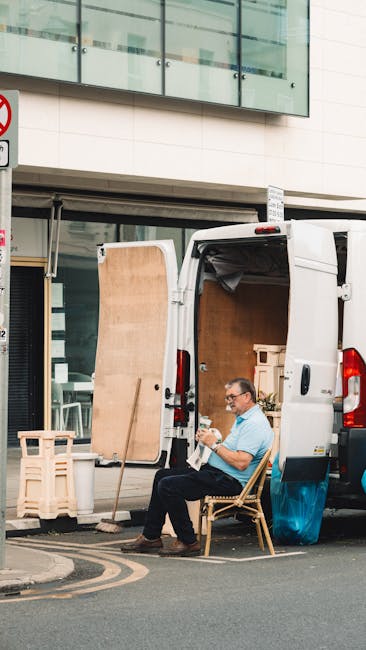 A man sitting on a wooden chair outside a residential building during a home relocation process, with the back doors of a white moving van open revealing large cardboard boxes and wrapped furniture inside. Nearby, a wooden panel is propped against the van, and a small folding stool is visible on the pavement. The man is wearing casual clothing, including a light blue shirt and dark trousers, and appears to be reviewing a document or instructions. The scene is set in an urban environment with a modern glass-fronted building above, and the area is prepared for furniture transport as part of packing and loading activities handled by Hammersmith Removals, a professional removals company. Equipment such as straps or blankets is not visible, but the focus on boxes and furniture supports processes involved in house removals, especially for small moves near Hammersmith Broadway W6.