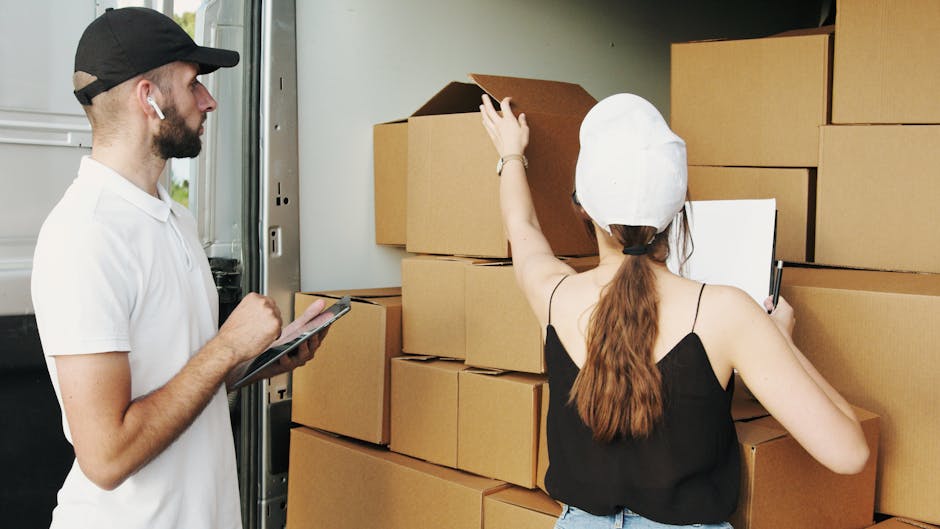 A man dressed in a white polo shirt and a black cap is standing indoors, holding a clipboard and pen, appearing to be taking notes or inspecting. Behind him, there are numerous cardboard boxes stacked on top of each other, some with visible packing tape, indicating an ongoing packing or loading process for a home relocation. The setting features a door or a large opening leading to a storage or loading area, with natural light illuminating the scene. The boxes vary in size and are arranged in a neat stack, suggesting organized packing for a small move. This image reflects the professional packing and furniture transport activities associated with house removals and moving services provided by Hammersmith Removals, supporting the topic of small move logistics and packing preparations.