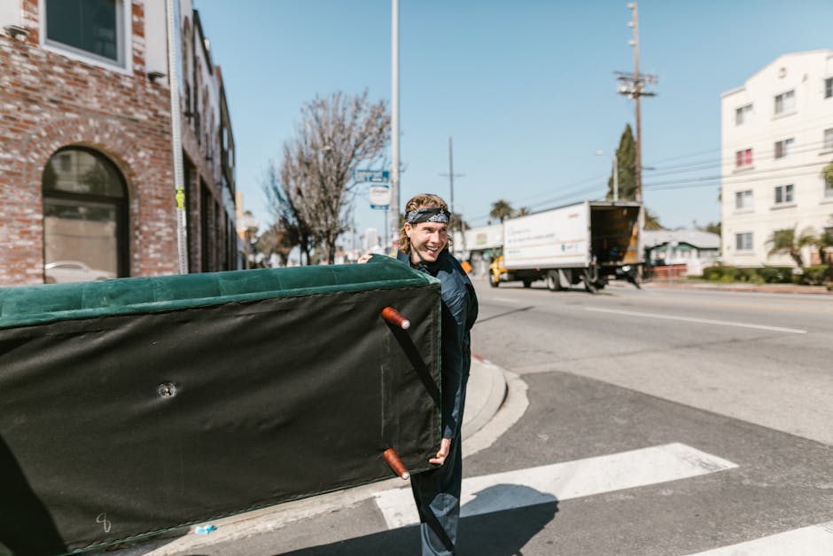 A person with light brown hair, wearing a headband and a dark jacket, is standing outside on a city street, carrying a large black moving pad or blanket with orange handles. The individual appears to be involved in a home relocation process as part of furniture transport or packing and moving activities facilitated by Hammersmith Removals, visible from the context of a moving van and boxes nearby. The street features a brick building with arched windows on the left and a traffic lane with vehicles, including a yellow and white truck, in the background. The environment is bright and clear, with a tree, power lines, and residential or commercial buildings in view, capturing a typical urban moving scene that supports house removals and relocation logistics.