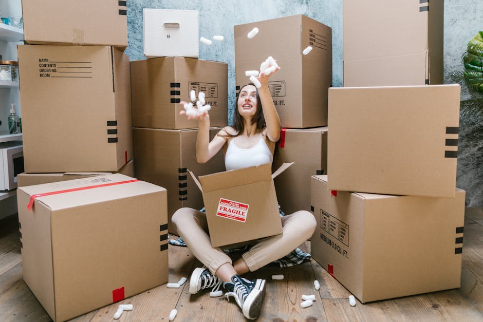 A woman sitting cross-legged on a hardwood floor surrounded by numerous cardboard moving boxes of various sizes, some sealed with red packing tape and others open, revealing packing materials. The woman is smiling and tossing white packing peanuts into the air while sitting inside one open box, which has a red 'fragile' label on its side. Behind her, additional boxes are stacked against a textured wall with a visible doorway, indicating an indoor setting. Equipment such as a trolley or straps may be nearby, used for the furniture transport and home relocation process. This scene captures the packing and loading stages of a house removal conducted by Hammersmith Removals, illustrating the preparation involved in furniture transport and moving logistics in a professional manner, aligned with the guidance found on the Hammersmith W6 King Street moving and removals guide page.
