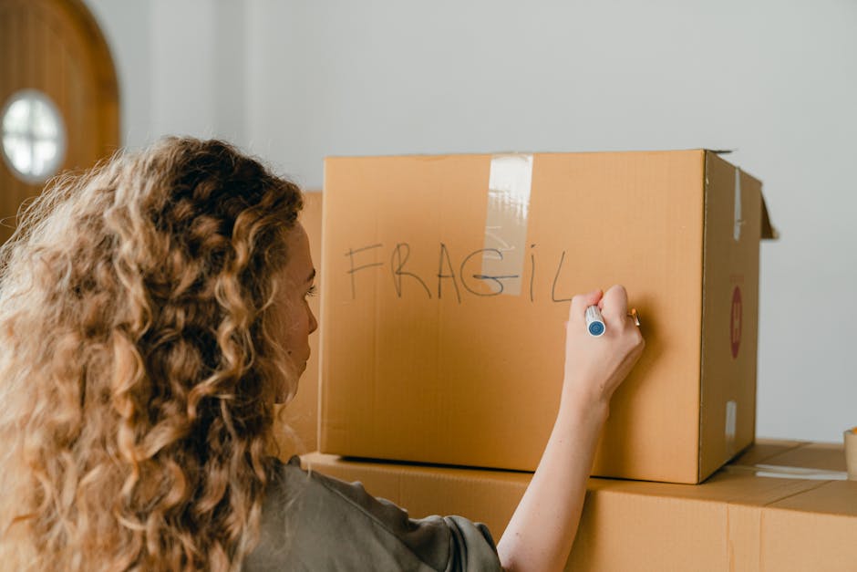 A woman with long, curly blonde hair is seen writing on a cardboard box with a blue marker. The box, labeled 'FRAGILE' in large letters, is positioned on top of other packed boxes in a well-lit indoor space, likely within a home. The woman has her back partially turned towards the camera, and her focus is on the box she is marking. The scene captures aspects of packing and preparation for home relocation, with cardboard boxes sealed with tape suggesting they are ready for transport. The environment appears tidy, and the lighting highlights the packing process, which is part of furniture transport and moving logistics managed by Hammersmith Removals. The setting includes a visible door to the left with a circular window, and the background wall is neutral in tone, emphasizing the focus on the packing activity related to home removals.
