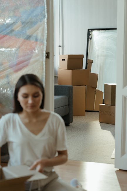 Inside a residential property, a woman with dark hair and wearing a white top is seated on the floor, focused on packing or unpacking items from a cardboard box. The room features a light-colored carpet and a partially open white door. In the background, several cardboard moving boxes of various sizes are stacked near a full-length mirror leaning against the wall, with additional boxes on the floor. To the left, a portion of a blue plastic-covered object or furniture is visible, indicating ongoing packing or home relocation activities. A grey sofa is also partially visible, situated against the wall beneath a large window or mirror. The scene is well-lit with natural light, illustrating a typical home moving process managed by a professional removal service, such as Hammersmith Removals, supporting home relocation and furniture transport tasks.