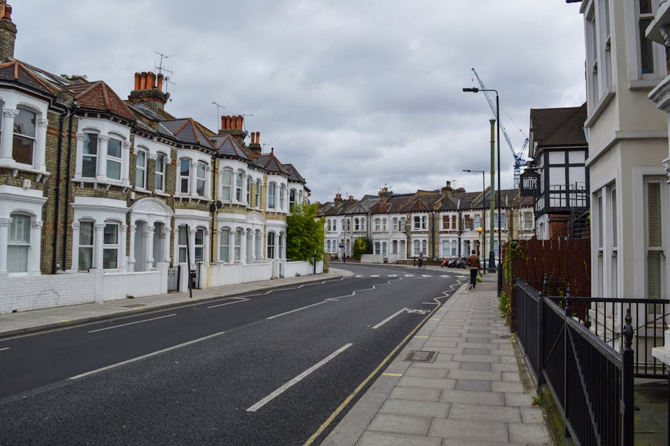 A quiet residential street with Victorian-style terraced houses featuring white facades, bay windows, and red tiled roofs, under a cloudy sky. The pavement on the right side of the image is bordered by black metal railings, with some plants growing along the fence. A pedestrian is walking along the sidewalk, and a blue crane is visible in the background, indicating ongoing construction. The street is lined with parked cars and has a traffic lane with marked lines, with no moving vehicles present. Visible street lighting and bollards are also part of the scene. This setting reflects a typical established neighbourhood suitable for house removals and home relocation services, like those offered by Hammersmith Removals, with a focus on transportation and packing logistics relevant to the process of moving homes in the area.