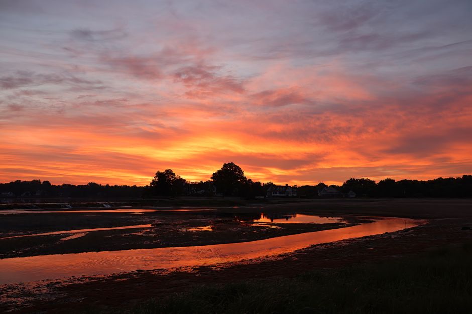 A scenic sunset over a riverbank with vibrant orange, pink, and purple hues in the sky, reflected on the calm water below. Silhouettes of trees line the horizon, and a few houses are visible in the distance. The natural outdoor environment creates a peaceful and picturesque scene. This image does not depict any furniture, boxes, appliances, or moving equipment related to house removals or relocation services, and no physical actions or transport activities are visible. It visually emphasizes a serene outdoor setting, which may be used as a background or contextual imagery for content about home relocation or moving logistics, such as those discussed by Hammersmith Removals on their Stamford Brook house removals page.